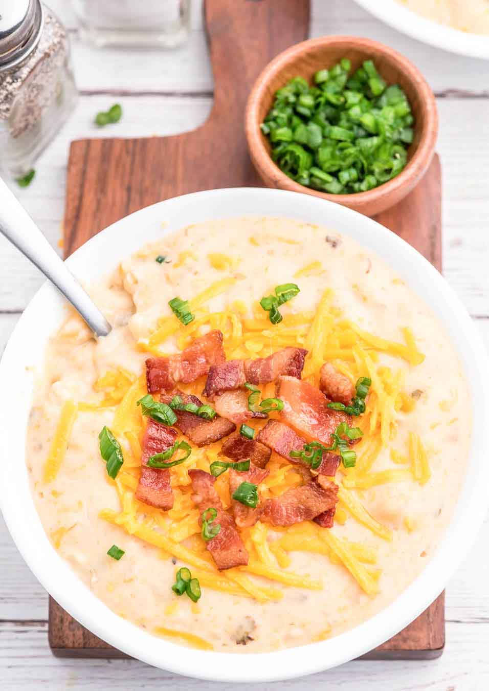 bowl of loaded potato soup with chopped bacon, sliced green onions and shredded cheese garnish being served on a small wooden board with a handle. Slow Cooker Baked Potato Soup